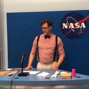 A man (Steven) in a redish pink plaid shirt with a black bow tie and suspenders standing at a blue podium with a microphone in front of a blue wall with a NASA logo