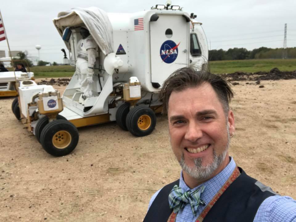 A selfie shot of Steven in a blue plaid shirt with blue vest and plaid bowtie in front of a white NASA rover vehicle