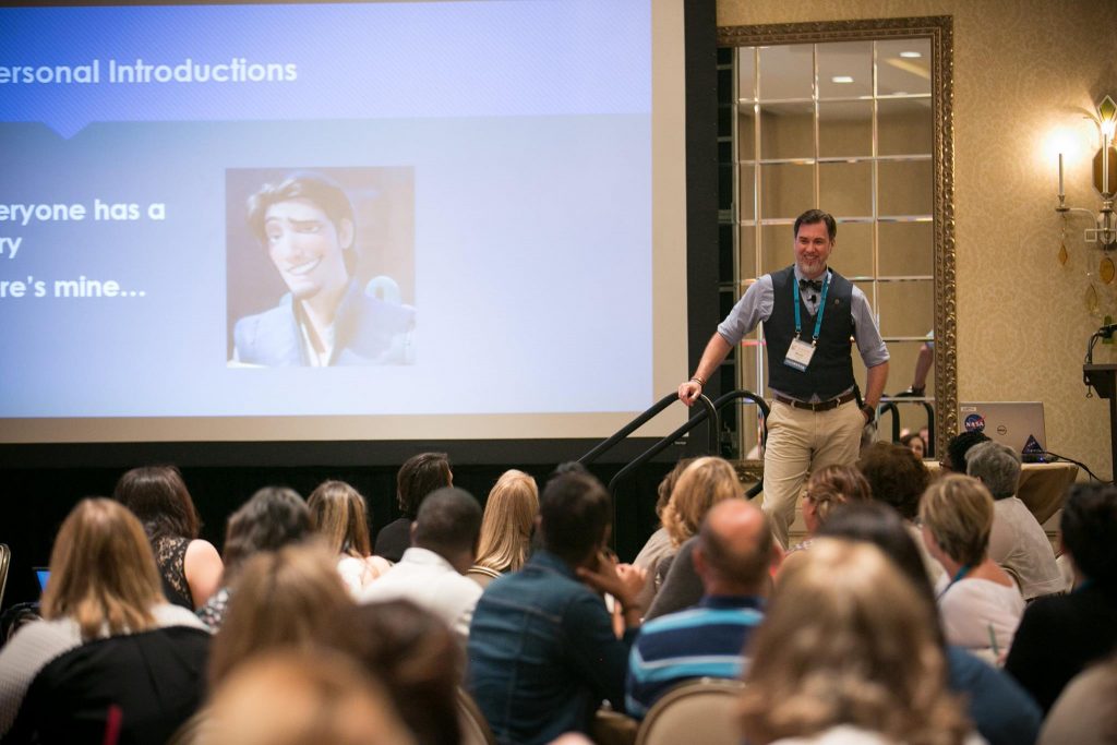 Steven standing wearing khakis, blue shirt, and blue vest next to a projector screen partially showing a slide titled "Personal Introductions". The back of audience members heads is in the foreground.