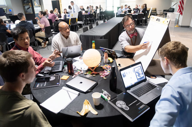 Five people sitting around a table with a black table cloth, Steven is holding a big sheet of paper writing, others have their laptops open in front of them.