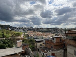 View of Belo Horizonte, Brazil showing buildings and a cloudy sky. In the foreground there are concrete and block homes with aluminum roofs. In the background, there are rows of newer beige and salmon-colored apartment blocks (approximately 4-7 floors tall). A greenspace is visible on the left.