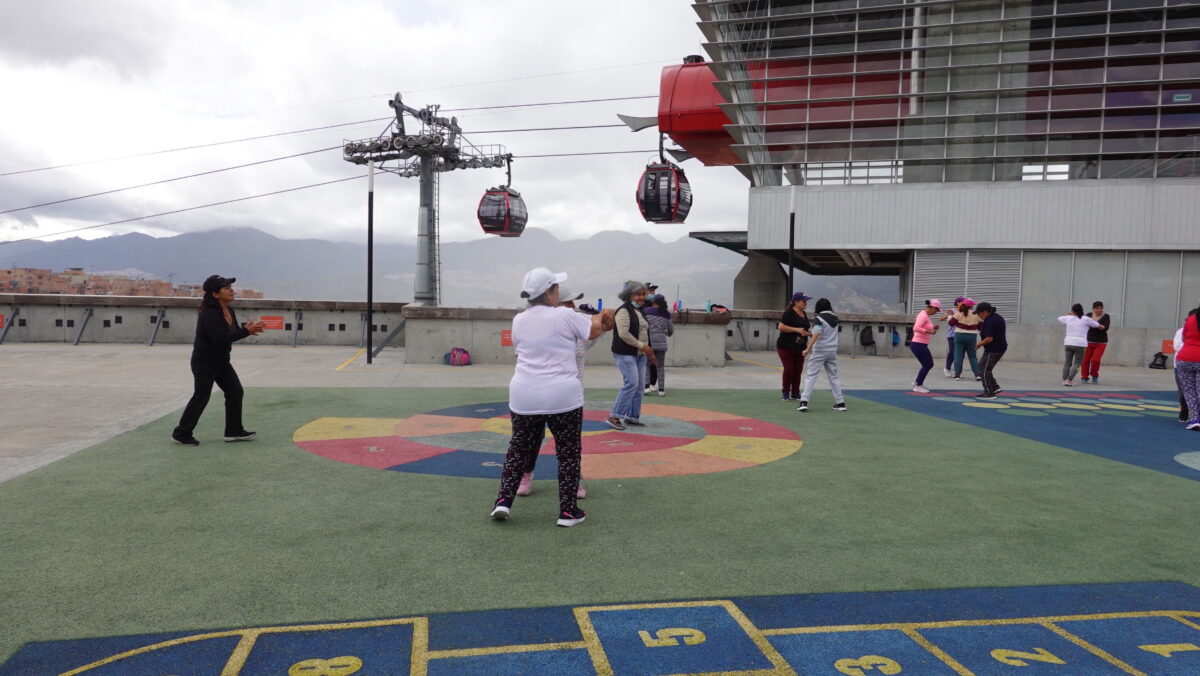 Women exercising on a rooftop with a colorful surface as part of the Blocks of Care project in Bogotá, Colombia. Cable cars are visible in the background.